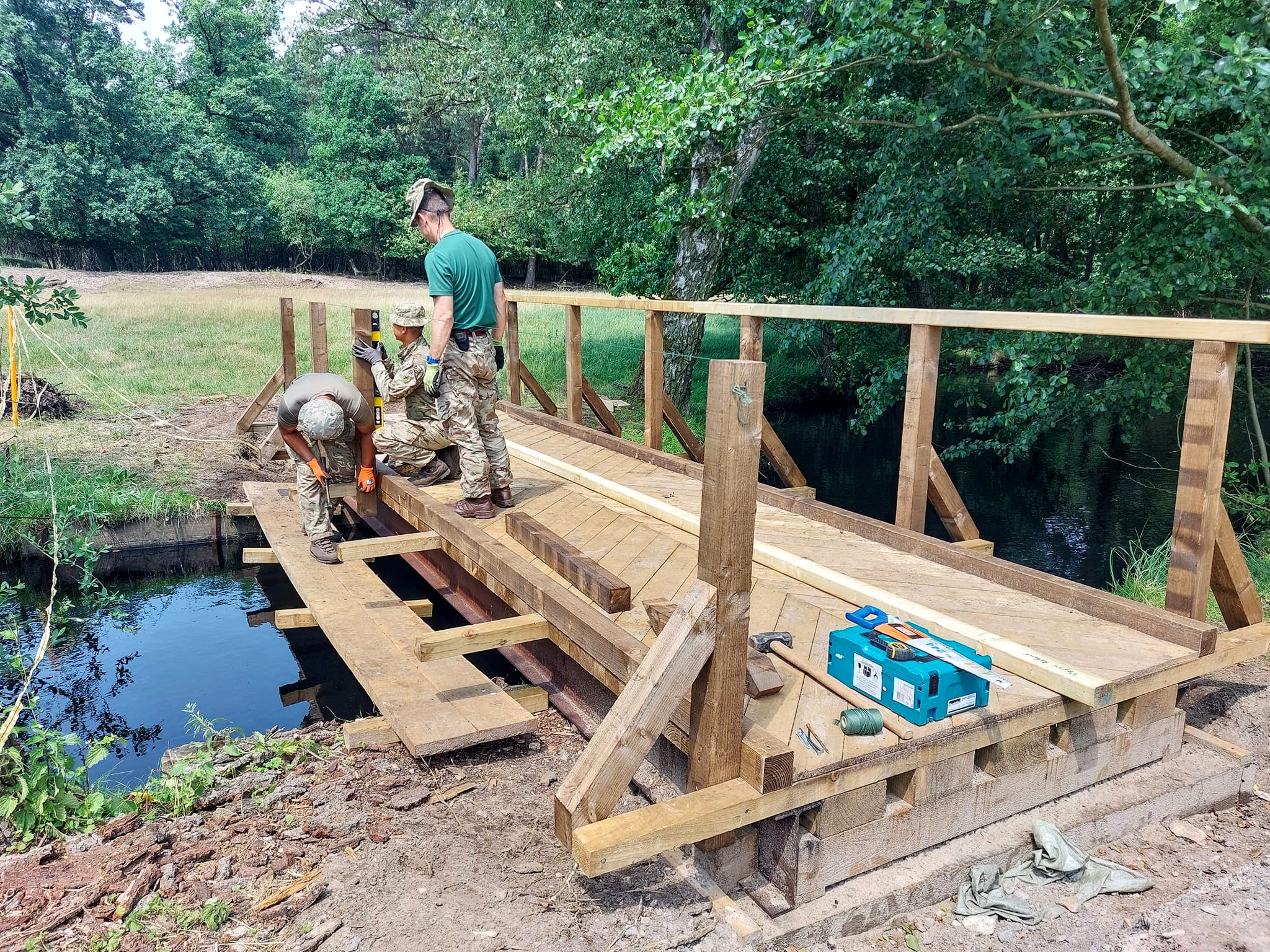 A team from 102 Field Squadron standing on and working alongside a wooden bridge under construction across a small stream.