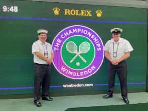 Two Reservists from HMS Dalriada standing either side of a Wimbledon Championships logo.