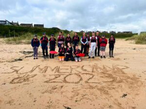 A group of Sea Cadets posing together on the beach, brandishing a kayaking oar and the words Summer Camp 2023 written in the sand.