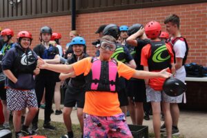 A group of Army Cadets from West Lowland Battalion preparing for water sports, fronted by a Cadet in large swimming goggles, an orange t-shirt and a pink life jacket, posing dramatically.