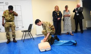 A male Cadet kneels in front of a Little Anne CPR dummy to begin First Aid practice, guided by the Instructor standing to his left and observed by the Lord Provost who stands behind him. Also behind the Cadet are the Chief and Deputy Chief Executives of Lowland RFCA.