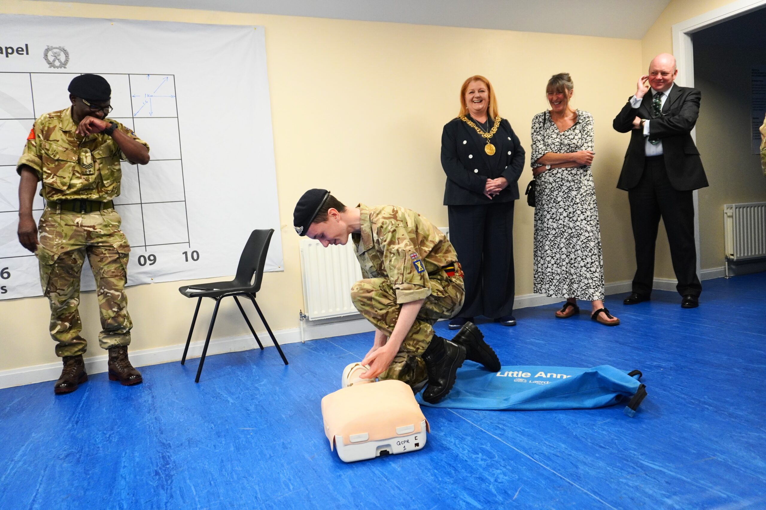 A male Cadet kneels in front of a Little Anne CPR dummy to begin First Aid practice, guided by the Instructor standing to his left and observed by the Lord Provost who stands behind him. Also behind the Cadet are the Chief and Deputy Chief Executives of Lowland RFCA.