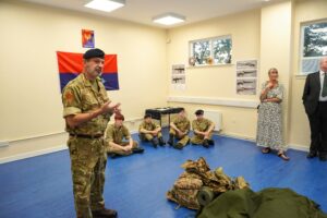 A Cadet Force Adult Volunteer talks Cadets through the equipment they need for effective outdoor survival. In the right corner, a group of Cadets are seated on the floor and listening to the instructor.