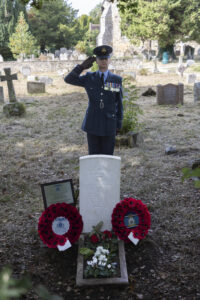 Squadron Leader Read salutes at the grave of Flight Lieutenant Freddie Rushmer, with poppy wreaths laid either side of the white headstone and a commemorative plaque for Jean Liddicoat on the left hand side.